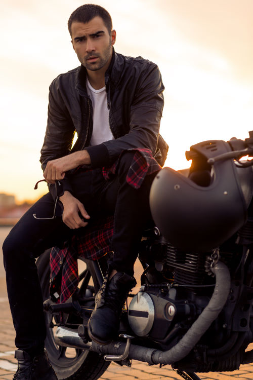 Handsome rider guy with beard and mustache in black leather biker jacket sit on classic style cafe racer motorcycle rooftop at sunset. Bike custom made in vintage garage. Brutal fun urban lifestyle.